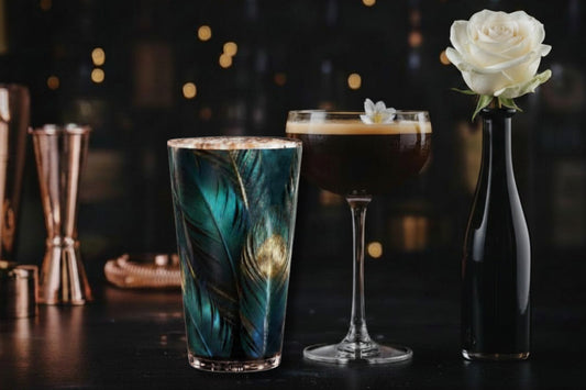 Glas mit türkis-goldenem Federmotiv in stilvoll dekorierter Bar mit weiterem Coctailglas und weißer Rose in schwarzer Vase mit stimmungsvoller Beleuchtung