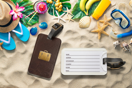 Kofferanhänger mit goldenem Reisekoffer-Motiv auf brauner Lederoptik am Strand mit geöffneter Rückseite und Kartenfach, umgeben von sommerlichen Reiseaccessoires.