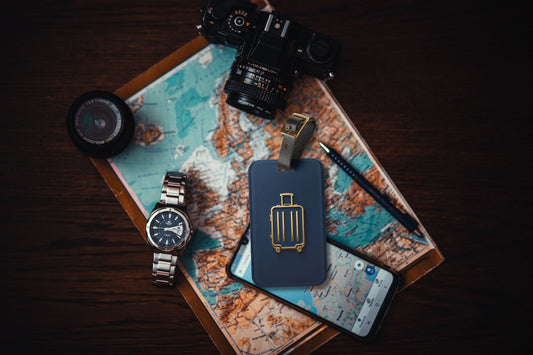 Kofferanhänger mit goldenem Koffersymbol auf tiefblauem Hintergrund auf einer Weltkarte arrangiert, umgeben von Kamera, Armbanduhr, Smartphone und Reiseutensilien.