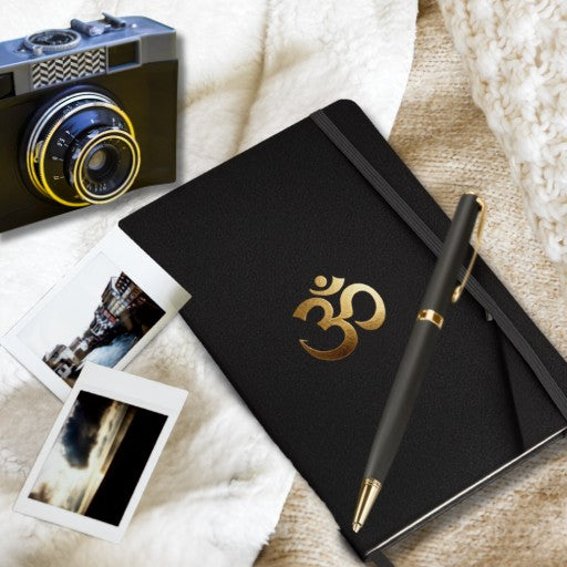 Hardcover Notizbuch mit goldenem Om-Symbol auf schwarzem Einband, dekoriert mit Kamera, Sofortbildern und Stift in einer gemütlichen Lifestyle-Szene.