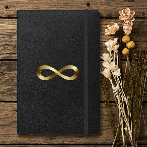 Hardcover Notizbuch mit Unendlichkeitszeichen in Goldoptik auf schwarzem Einband, dekoriert mit Trockenblumen auf einem rustikalen Holzhintergrund.