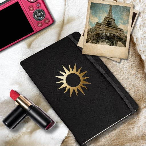Spiralnotizbuch mit goldenem Sonnensymbol auf schwarzem Einband, dekoriert mit Kamera, Lippenstift und Vintage-Fotografien in einer stilvollen Lifestyle-Szene.
