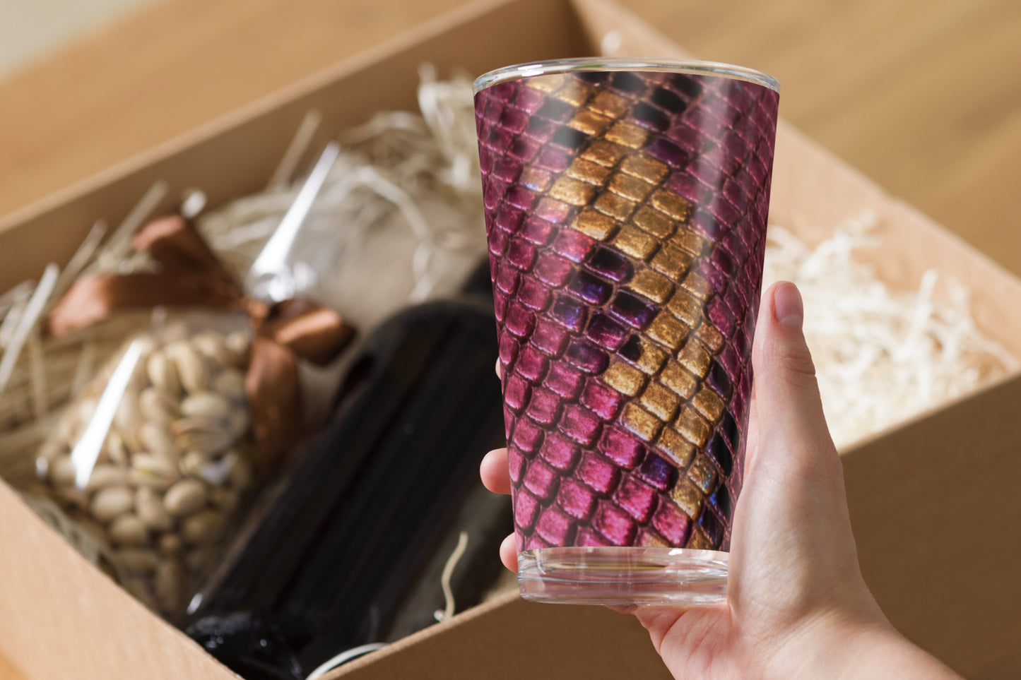 Handaufnahme der Vorderansicht des luxuriösen Magenta-Gold Glases mit strukturierter Reptilienoptik neben stilvoller Versandverpackung