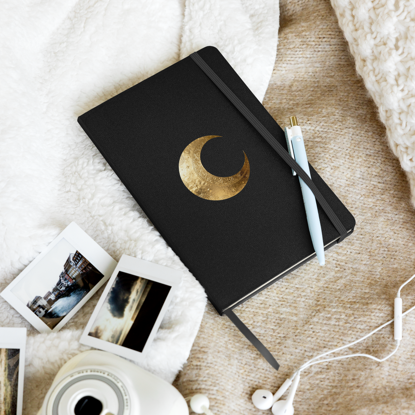 Hardcover Notizbuch mit goldenem Mondsymbol auf schwarzem Einband, dekorativ mit Stift, Fotos und Kaffeetasse arrangiert.