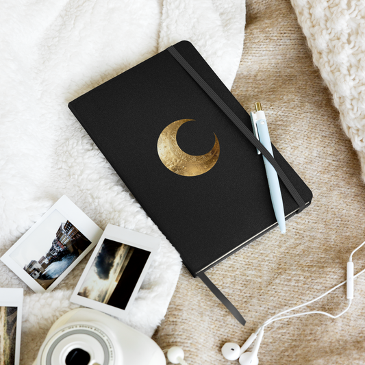 Hardcover Notizbuch mit goldenem Mondsymbol auf schwarzem Einband, dekorativ mit Stift, Fotos und Kaffeetasse arrangiert.