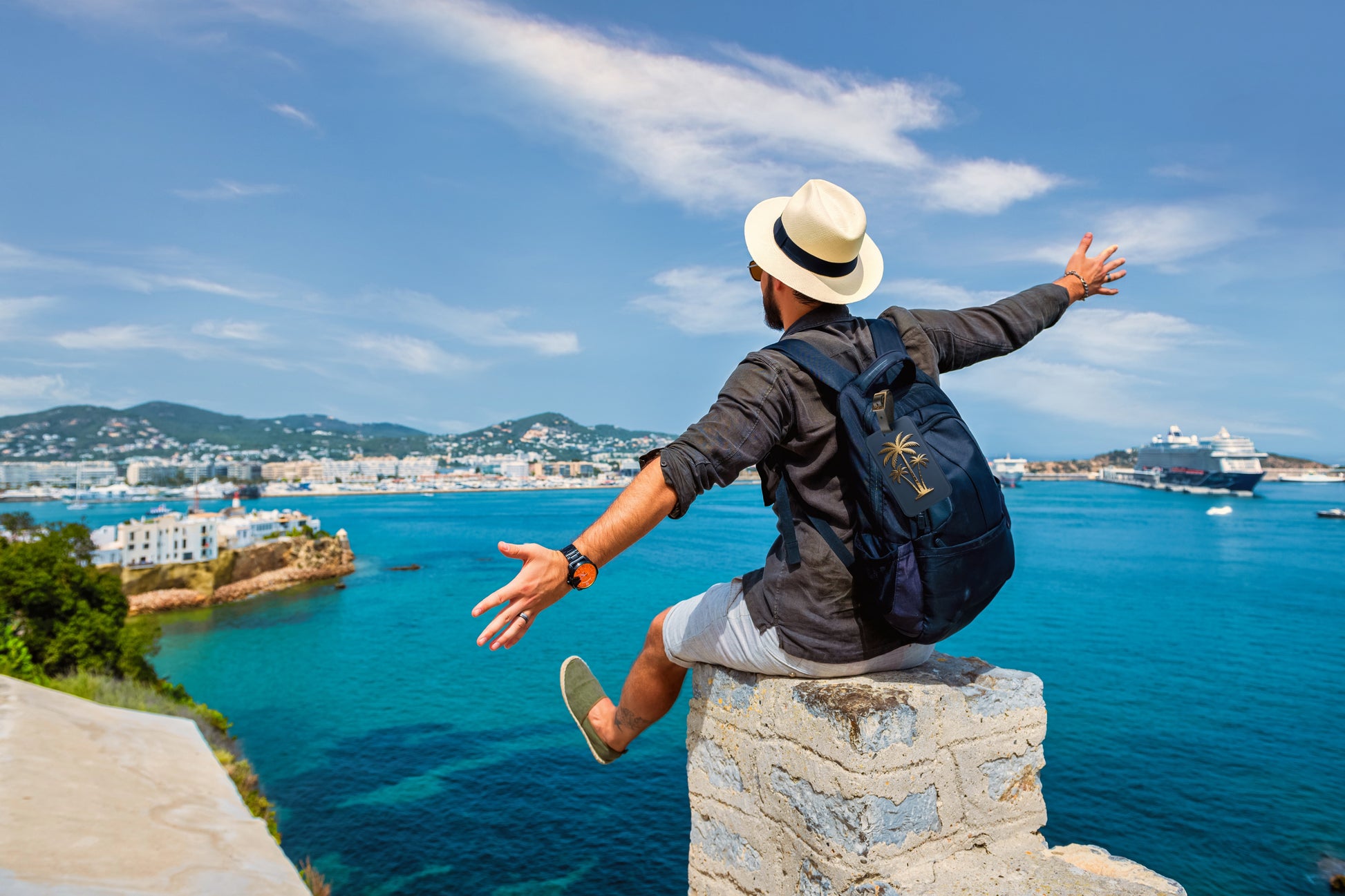 Kofferanhänger mit goldener Palmensilhouette auf tiefblauem Hintergrund befestigt an einem Rucksack vor einer Küstenlandschaft mit türkisblauem Meer und Urlaubsatmosphäre.