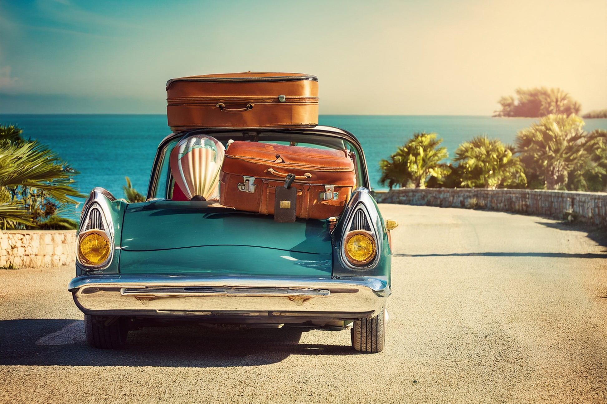 Kofferanhänger mit goldenem Reisekoffer-Motiv auf brauner Lederoptik vor einem klassischen Cabrio mit aufgestapeltem Vintage-Reisegepäck an einer Küstenstraße.