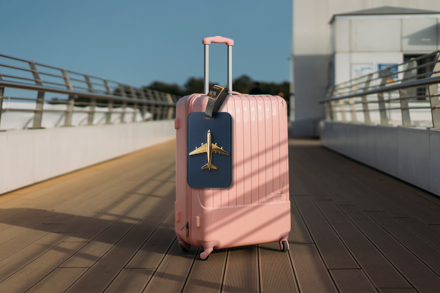 Kofferanhänger mit goldenem Flugzeug auf tiefblauem Hintergrund befestigt an einem rosafarbenen Hartschalenkoffer auf einer modernen Reisepromenade.