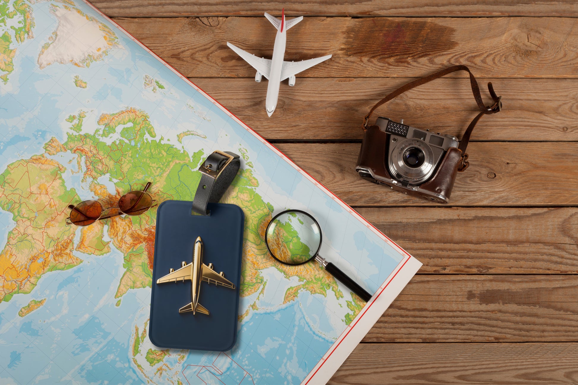 Kofferanhänger mit goldenem Flugzeug auf tiefblauem Hintergrund auf einer Weltkarte arrangiert, umgeben von Reisekamera, Lupe und Flugzeugmodell als Symbol für Fernreisen und Entdeckungen.