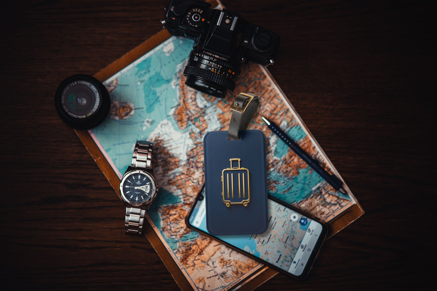Kofferanhänger mit goldenem Koffersymbol auf tiefblauem Hintergrund auf einer Weltkarte arrangiert, umgeben von Kamera, Armbanduhr, Smartphone und Reiseutensilien.