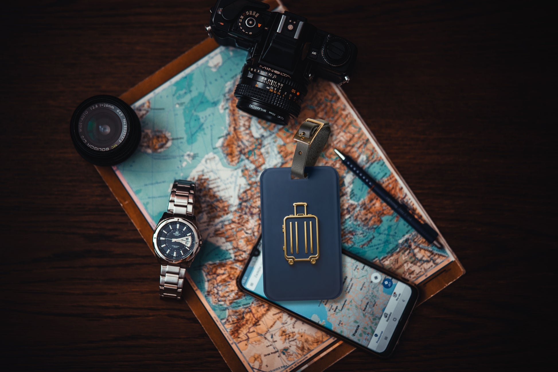 Kofferanhänger mit goldenem Koffersymbol auf tiefblauem Hintergrund auf einer Weltkarte arrangiert, umgeben von Kamera, Armbanduhr, Smartphone und Reiseutensilien.