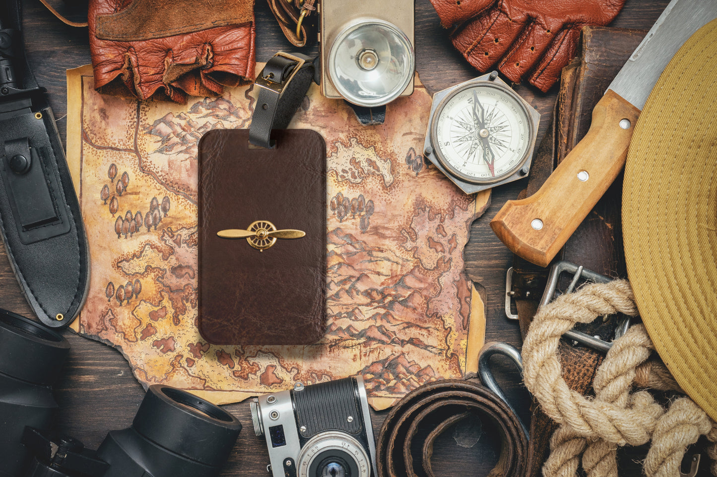 Kofferanhänger mit goldenem Propellerflugzeug auf dunkelbrauner Lederoptik, arrangiert auf einer historischen Weltkarte mit Kompass und Reiseutensilien im Vintage-Stil.