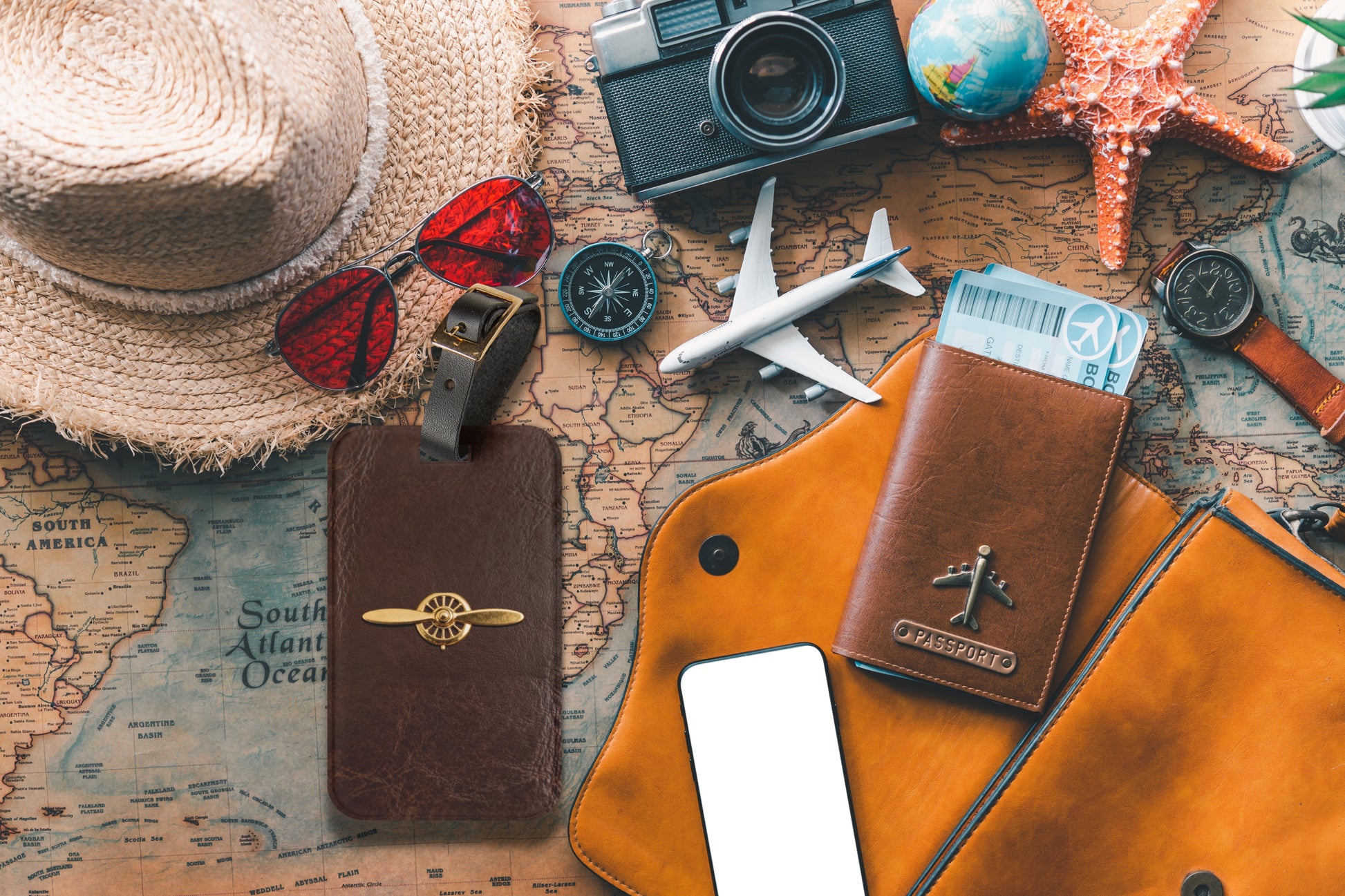 Kofferanhänger mit goldenem Propellerflugzeug auf dunkelbrauner Lederoptik, präsentiert zwischen Reisepasshülle, Weltkarte, Kamera, Sonnenbrille und weiteren Reiseaccessoires.